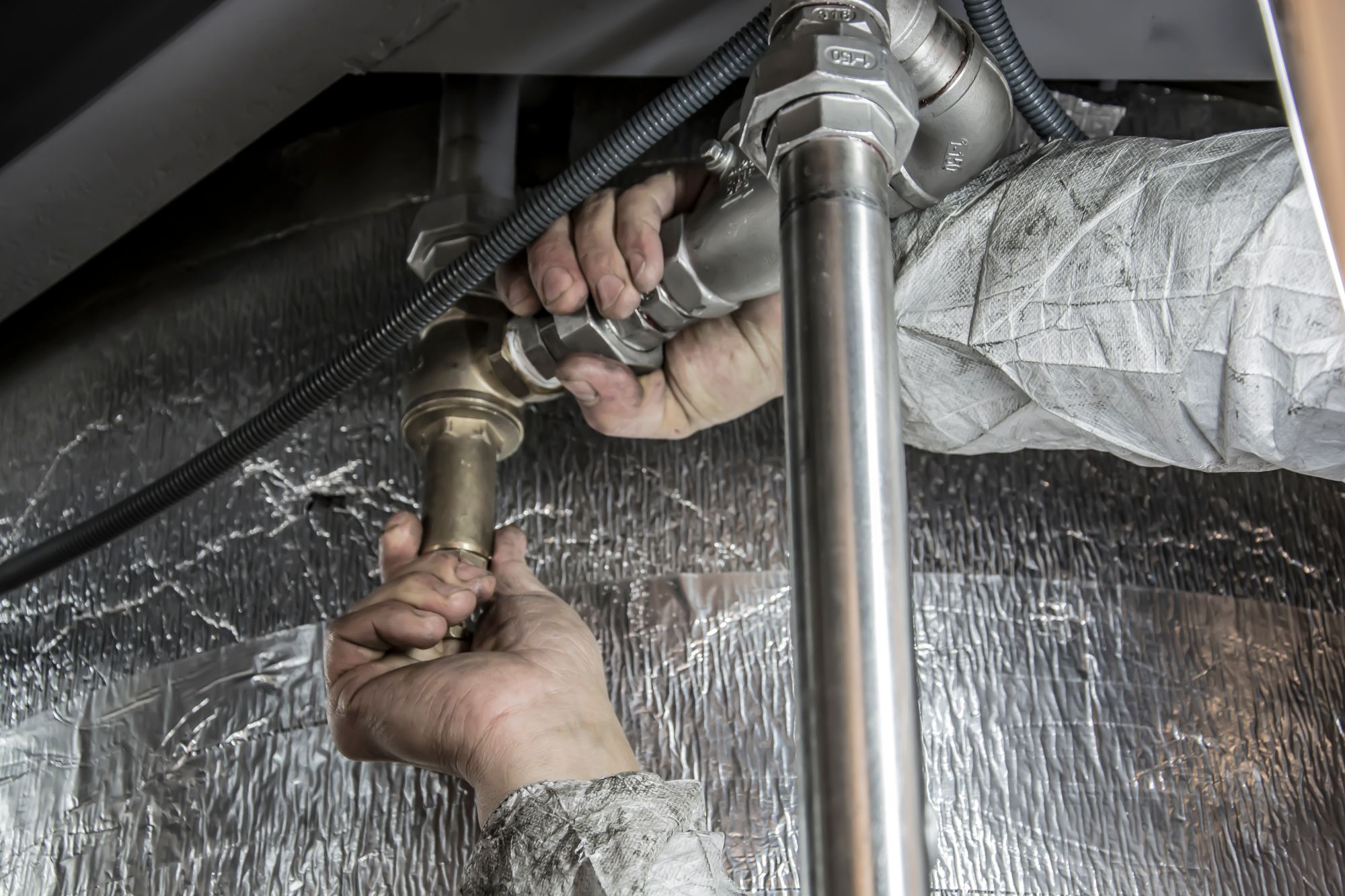 A plumber's grease-stained hands threading pipe fittings against foil insulation